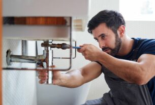 Encanador realizando manutenção no encanamento sob a pia, ajustando tubulação e conexões hidráulicas com ferramenta profissional.