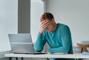 Homem sentado à mesa com notebook aberto, apoiando a mão na cabeça em sinal de preocupação enquanto trabalha em ambiente interno iluminado por janela.