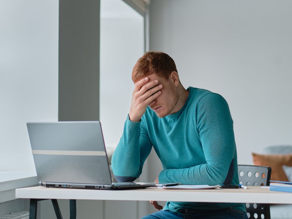 Homem sentado à mesa com notebook aberto, apoiando a mão na cabeça em sinal de preocupação enquanto trabalha em ambiente interno iluminado por janela.
