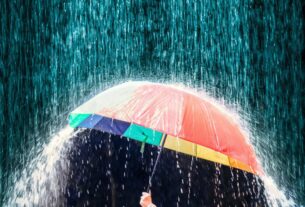 Guarda-chuva colorido aberto sob chuva intensa, com água escorrendo pelas laterais e gotas caindo em grande volume ao fundo escuro.