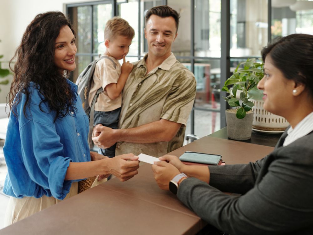 Família sendo atendida na recepção de empresa, realizando cadastro com a recepcionista em balcão de atendimento organizado.