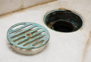 Ralo de banheiro com tampa metálica removida, mostrando abertura do encanamento no piso cerâmico e possível causa de cheiro ruim no ralo.