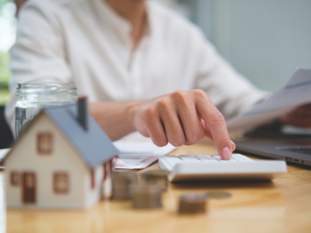 Pessoa calculando despesas com calculadora ao lado de uma miniatura de casa e moedas empilhadas sobre a mesa, representando cálculo de IPTU e custos imobiliários.
