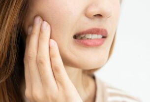 Mulher com expressão de desconforto tocando a bochecha, indicando dor de dente ou inflamação na região da mandíbula.