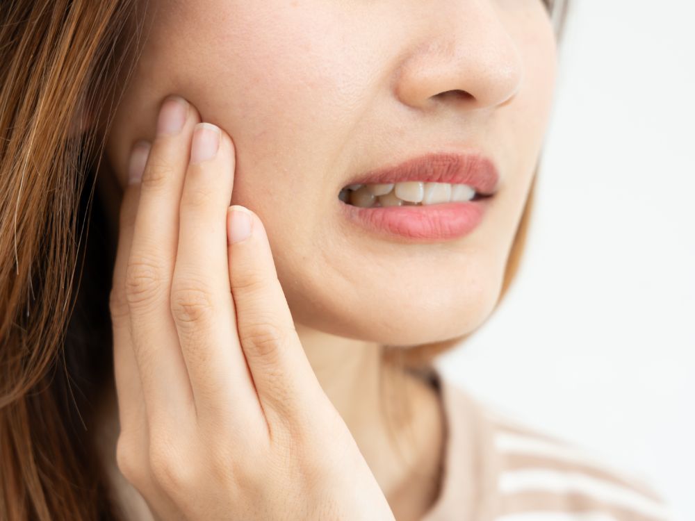 Mulher com expressão de desconforto tocando a bochecha, indicando dor de dente ou inflamação na região da mandíbula.