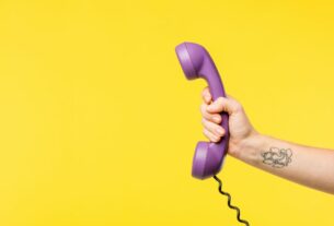 Mão segurando telefone roxo com fio sobre fundo amarelo, representando contato e atendimento pelo telefone da prefeitura de Volta Redonda.
