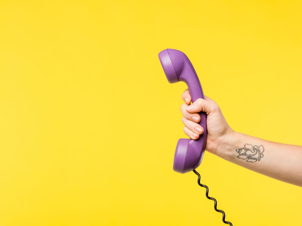 Mão segurando telefone roxo com fio sobre fundo amarelo, representando contato e atendimento pelo telefone da prefeitura de Volta Redonda.