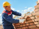 Pedreiro realizando assentamento de tijolos em obra de alvenaria, utilizando equipamentos de proteção durante construção residencial.