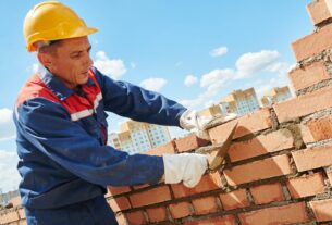 Pedreiro realizando assentamento de tijolos em obra de alvenaria, utilizando equipamentos de proteção durante construção residencial.
