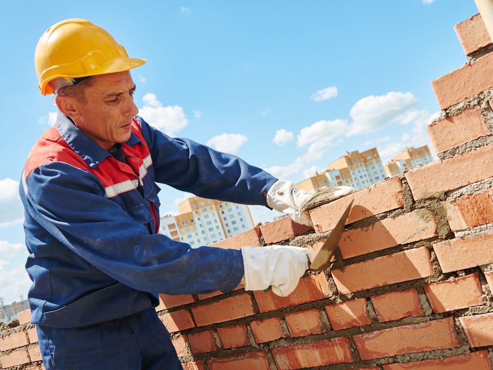 Pedreiro realizando assentamento de tijolos em obra de alvenaria, utilizando equipamentos de proteção durante construção residencial.